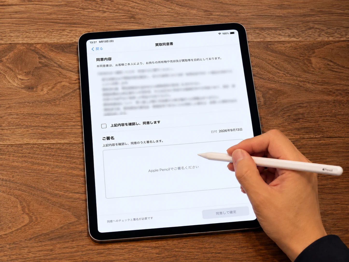 木の天板に置かれたiPadの買取同意書と、Apple Pencilを持つ手。