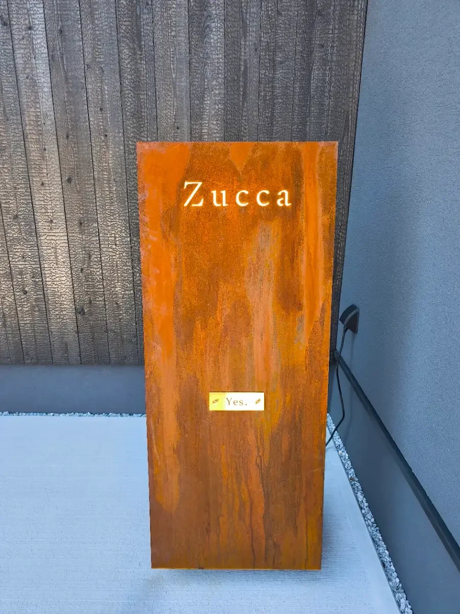 黒い外壁の前に立つ、Zuccaの文字とYES表示が光るコールテン鋼の看板。